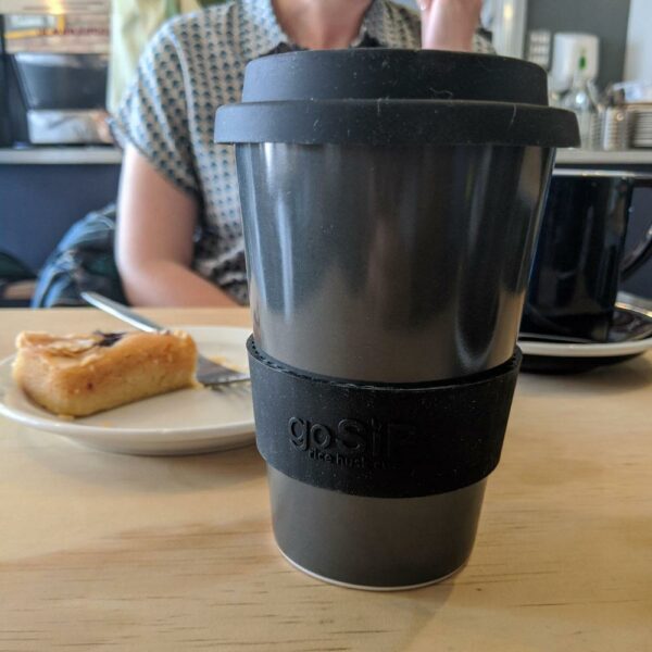 Reusable travel mug charcoal coloured on a table with a plate of food.