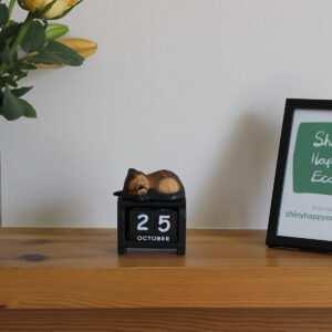 Front view of sleeping cat block calendar in dark brown on black pictured on a mantlepiece.