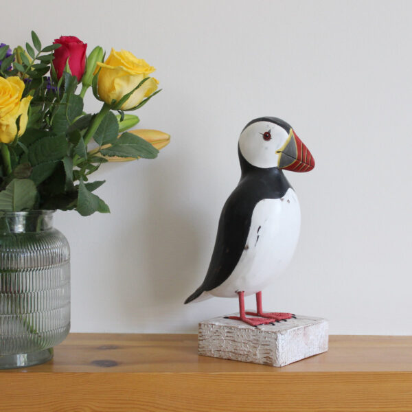 Wooden Puffin ornament on a mantlepiece next to flowers, hand made and painted in a rustic style and available from Shiny Happy Eco.