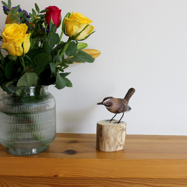 Hand carved and painted Wooden Wren Ornament on driftwood pictured on a mantlepiece next to a vase of flowers available from Shiny Happy Eco.